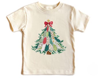 May include: Cream-colored short-sleeved t-shirt featuring a Christmas tree design. The tree is composed of various fabric patches in green, red, and floral patterns, topped with a red bow and gold star. A festive apparel item.