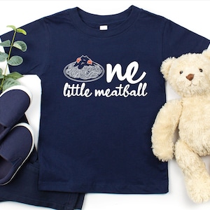 May include: Navy blue toddler t-shirt with the words "one little meatball" in white. The shirt features a spaghetti and meatball graphic. The image also includes navy blue shoes, pants, a teddy bear, and greenery.