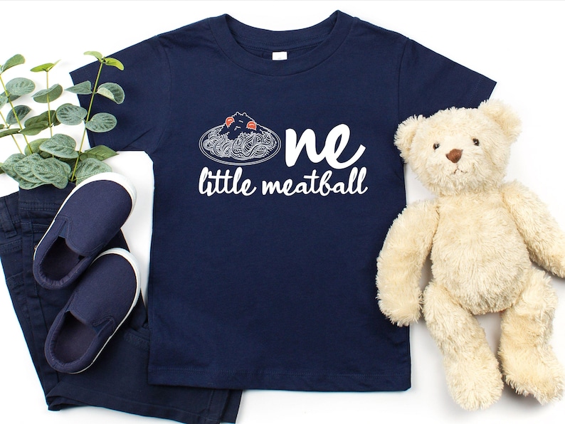 May include: Navy blue toddler t-shirt with the words "one little meatball" in white. The shirt features a spaghetti and meatball graphic. The image also includes navy blue shoes, pants, a teddy bear, and greenery.