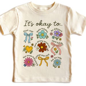 May include: Cream-colored t-shirt with the text "It's okay to..." and colorful illustrations. The design includes flowers, bows, and hearts, with phrases like "Start over" and "Ask for help". A cheerful and positive design.