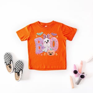 Puede incluir: Una camiseta naranja de manga corta con un estampado gr&aacute;fico de la palabra BOO en colores pastel, un fantasma gui&ntilde;ando un ojo con un lazo rosa de lunares, una calabaza sonriente, murci&eacute;lagos y telara&ntilde;as. El dise&ntilde;o incluye estrellas.