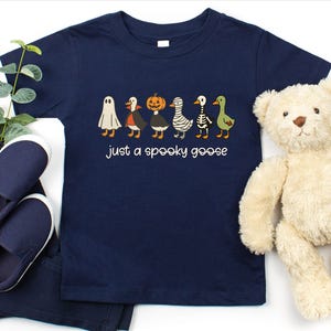 May include: Navy blue toddler t-shirt with the text "just a spooky goose." The shirt features a line of cartoon geese dressed in Halloween costumes, including a ghost, vampire, pumpkin, mummy, skeleton, and green goose. A pair of navy blue shoes and pants are next to the shirt.