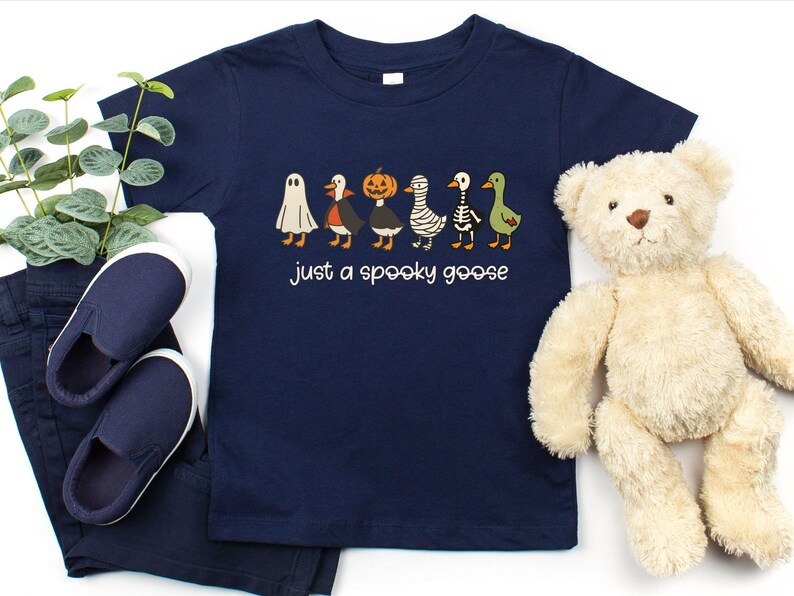 May include: Navy blue toddler t-shirt with the text "just a spooky goose." The shirt features a line of cartoon geese dressed in Halloween costumes, including a ghost, vampire, pumpkin, mummy, skeleton, and green goose. A pair of navy blue shoes and pants are next to the shirt.