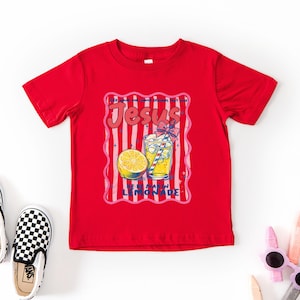 May include: A red t-shirt with a graphic that says "Jesus Loves Lemonade." The graphic includes a glass of lemonade, a lemon slice, and pink and white stripes. Black and white checkered slip-on shoes are on the left.
