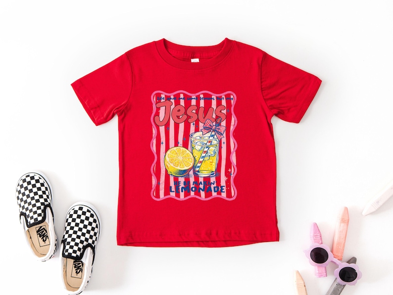 May include: A red t-shirt with a graphic that says "Jesus Loves Lemonade." The graphic includes a glass of lemonade, a lemon slice, and pink and white stripes. Black and white checkered slip-on shoes are on the left.