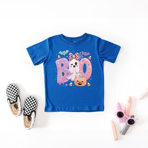 Puede incluir: Una camiseta azul real de manga corta con un estampado gr&aacute;fico de la palabra BOO en rosa y morado, un fantasma gui&ntilde;ando un ojo con un lazo rosa de lunares, una calabaza naranja sonriente, murci&eacute;lagos, telara&ntilde;as y caramelos de ma&iacute;z.