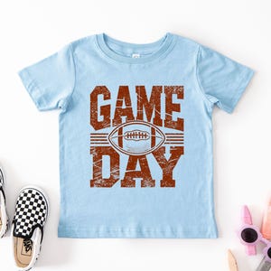 K&ouml;nnte beinhalten: Hellblaues T-Shirt mit dem Text "GAME DAY" in abgenutzten braunen Buchstaben und einer Football-Grafik. Das Bild zeigt ein Paar schwarz-wei&szlig; karierte Slip-On-Schuhe.