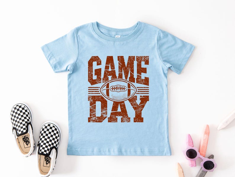 K&ouml;nnte beinhalten: Hellblaues T-Shirt mit dem Text "GAME DAY" in abgenutzten braunen Buchstaben und einer Football-Grafik. Das Bild zeigt ein Paar schwarz-wei&szlig; karierte Slip-On-Schuhe.
