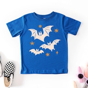 May include: A royal blue short-sleeve t-shirt featuring a graphic print of three cream-colored bats in flight, accented by several small, four-pointed orange stars scattered across the front of the garment.
