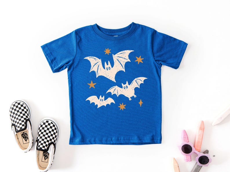 May include: A royal blue short-sleeve t-shirt featuring a graphic print of three cream-colored bats in flight, accented by several small, four-pointed orange stars scattered across the front of the garment.