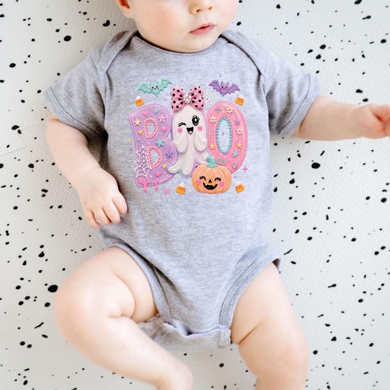 Puede incluir: Un body para beb&eacute; de color gris jaspeado con un estampado gr&aacute;fico de la palabra BOO en rosa y azul, un fantasma gui&ntilde;ando un ojo con un lazo rosa de lunares, una calabaza naranja sonriente, murci&eacute;lagos peque&ntilde;os y dulces en la parte frontal.