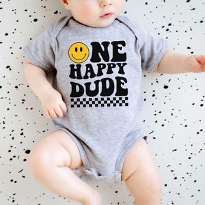 May include: A gray baby onesie with the text "ONE HAPPY DUDE" in black, a yellow smiley face, and a black and white checkered pattern. The onesie is short-sleeved.