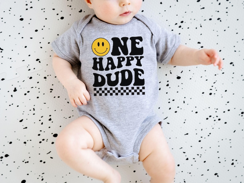 May include: A gray baby onesie with the text "ONE HAPPY DUDE" in black, a yellow smiley face, and a black and white checkered pattern. The onesie is short-sleeved.