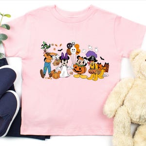 May include: A light pink t-shirt featuring a Halloween-themed graphic of Disney characters dressed in costumes. Goofy, Minnie Mouse, Mickey Mouse, and Pluto are depicted with pumpkins, candy, and balloons. The shirt is displayed with blue jeans, shoes, and a teddy bear.