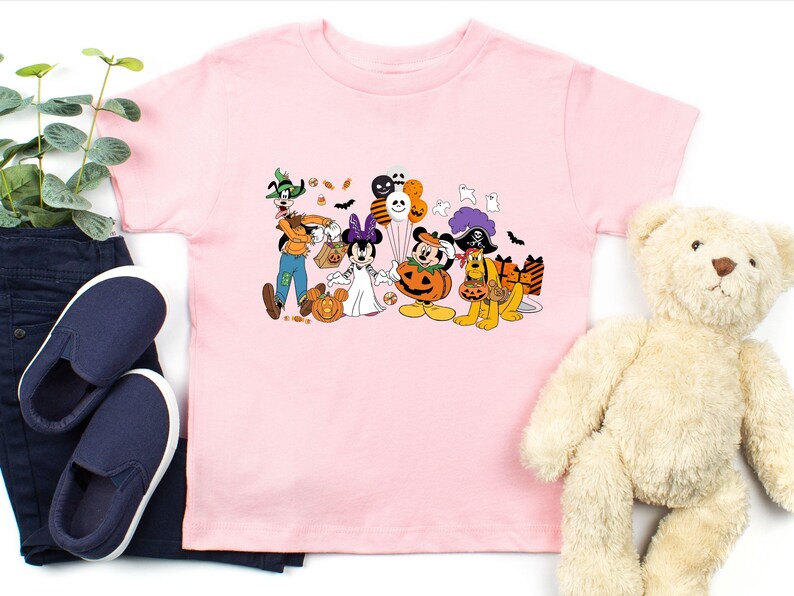 May include: A light pink t-shirt featuring a Halloween-themed graphic of Disney characters dressed in costumes. Goofy, Minnie Mouse, Mickey Mouse, and Pluto are depicted with pumpkins, candy, and balloons. The shirt is displayed with blue jeans, shoes, and a teddy bear.