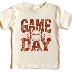 K&ouml;nnte beinhalten: Cremefarbenes Kurzarm-T-Shirt mit einem braunen Used-Look-Grafik. Die Grafik zeigt "GAME DAY" &uuml;ber und unter einer Football-Illustration. Das Shirt besteht aus einem weichen, bequemen Material.