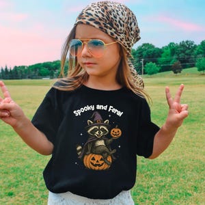 Peut inclure: T-shirt noir avec l'inscription "Spooky and Feral" et un raton laveur portant un chapeau de sorci&egrave;re, tenant une citrouille et assis sur une citrouille. La personne porte un foulard l&eacute;opard et des lunettes de soleil.