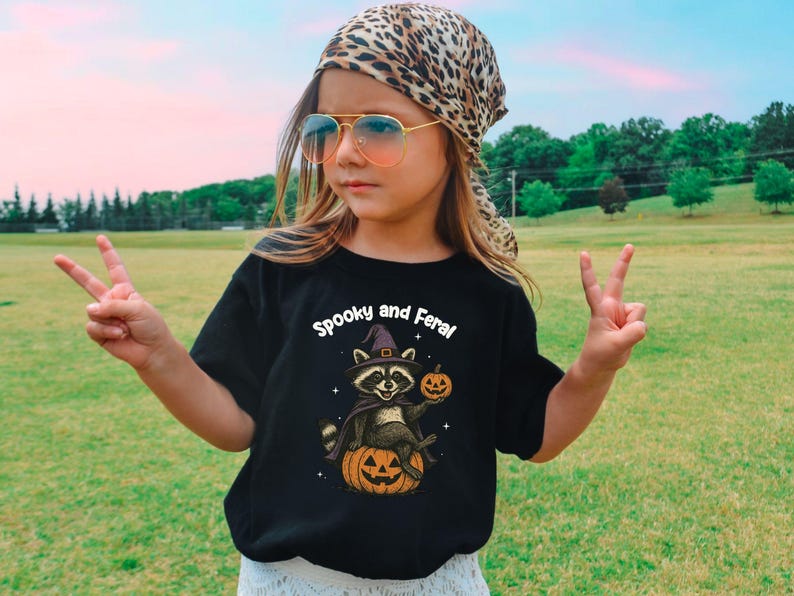 Peut inclure: T-shirt noir avec l'inscription "Spooky and Feral" et un raton laveur portant un chapeau de sorci&egrave;re, tenant une citrouille et assis sur une citrouille. La personne porte un foulard l&eacute;opard et des lunettes de soleil.