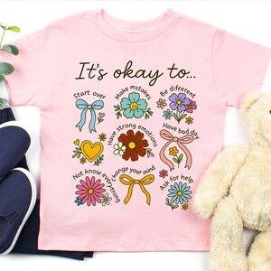 May include: Pink t-shirt with the text "It's okay to..." and colorful illustrations of flowers, bows, and hearts. The shirt is paired with navy blue slip-on shoes, dark blue pants, and a teddy bear.