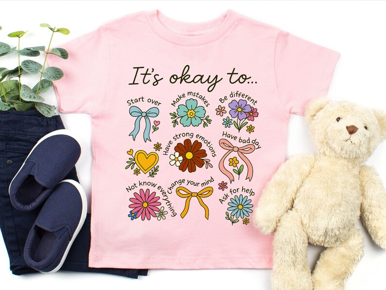 May include: Pink t-shirt with the text "It's okay to..." and colorful illustrations of flowers, bows, and hearts. The shirt is paired with navy blue slip-on shoes, dark blue pants, and a teddy bear.