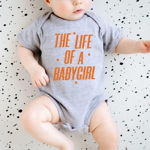 Pode incluir: Um body cinza para beb&eacute; com o texto "THE LIFE OF A BABYGIRL" em laranja. O texto est&aacute; centrado na frente do body. O beb&eacute; est&aacute; deitado numa superf&iacute;cie branca com salpicos pretos.