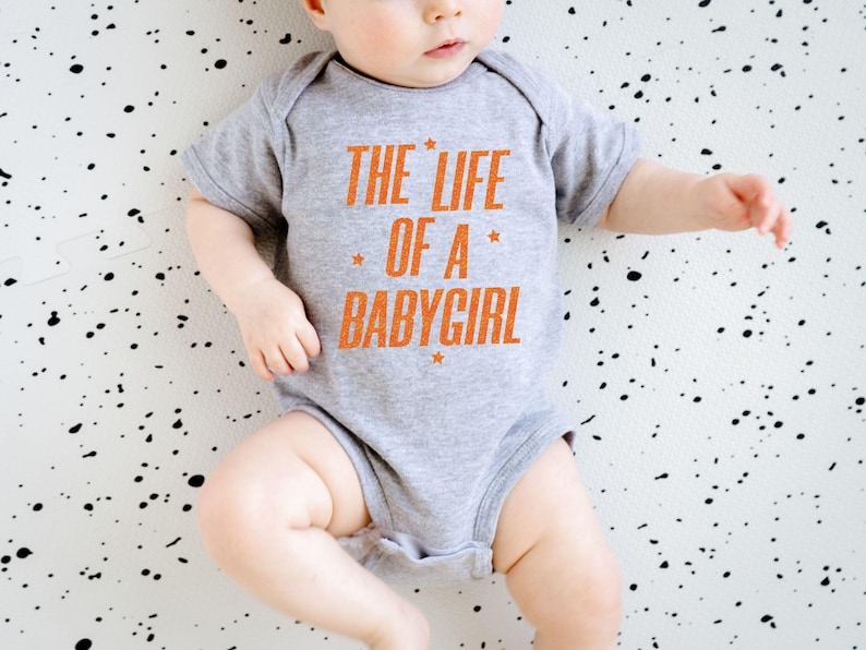 Pode incluir: Um body cinza para beb&eacute; com o texto "THE LIFE OF A BABYGIRL" em laranja. O texto est&aacute; centrado na frente do body. O beb&eacute; est&aacute; deitado numa superf&iacute;cie branca com salpicos pretos.