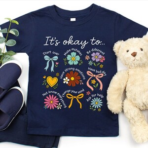 May include: Navy blue t-shirt with the text "It's okay to..." and colorful floral illustrations. The shirt includes the phrases "Start over", "Make mistakes", "Be different", "Have strong emotions", "Have bad day", "Not know everything", "Change your mind", and "Ask for help".