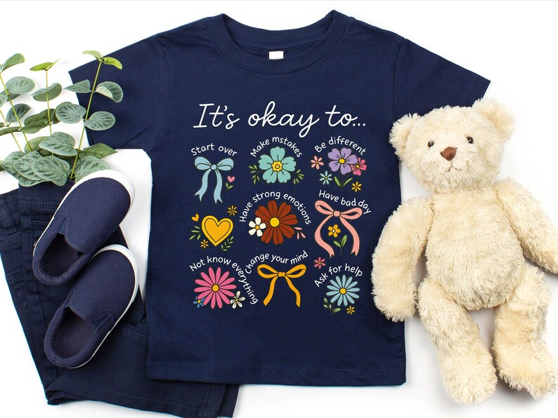 May include: Navy blue t-shirt with the text "It's okay to..." and colorful floral illustrations. The shirt includes the phrases "Start over", "Make mistakes", "Be different", "Have strong emotions", "Have bad day", "Not know everything", "Change your mind", and "Ask for help".