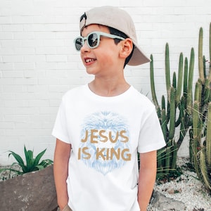 May include: A white t-shirt with the text "JESUS IS KING" in gold lettering and a blue lion design. The child is wearing a beige cap and light green sunglasses.