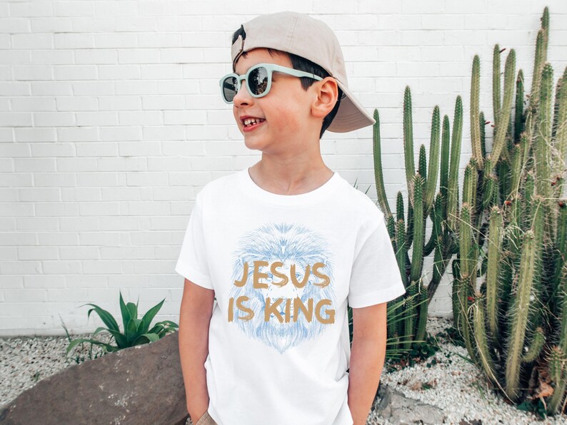 May include: A white t-shirt with the text "JESUS IS KING" in gold lettering and a blue lion design. The child is wearing a beige cap and light green sunglasses.