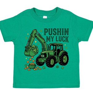 May include: A green toddler t-shirt featuring a green tractor with a bucket full of shamrocks and gold coins. The text "PUSHIN MY LUCK" is printed above the tractor. The design is in a cartoon style.