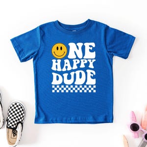 May include: A royal blue t-shirt with the words "ONE HAPPY DUDE" in white, featuring a yellow smiley face. The shirt has a checkered pattern at the bottom. Black and white checkered slip-on shoes are next to the shirt.