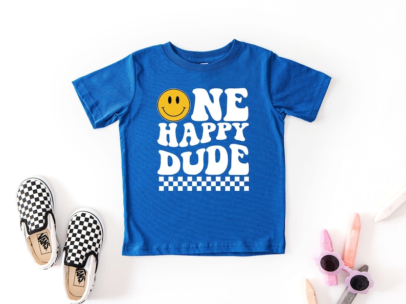 May include: A royal blue t-shirt with the words "ONE HAPPY DUDE" in white, featuring a yellow smiley face. The shirt has a checkered pattern at the bottom. Black and white checkered slip-on shoes are next to the shirt.