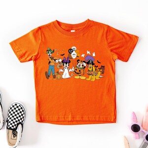 May include: Orange short-sleeved t-shirt featuring a Halloween-themed graphic with cartoon characters. The graphic includes characters dressed in costumes, pumpkins, and balloons. Black and white checkered slip-on shoes are next to the shirt.