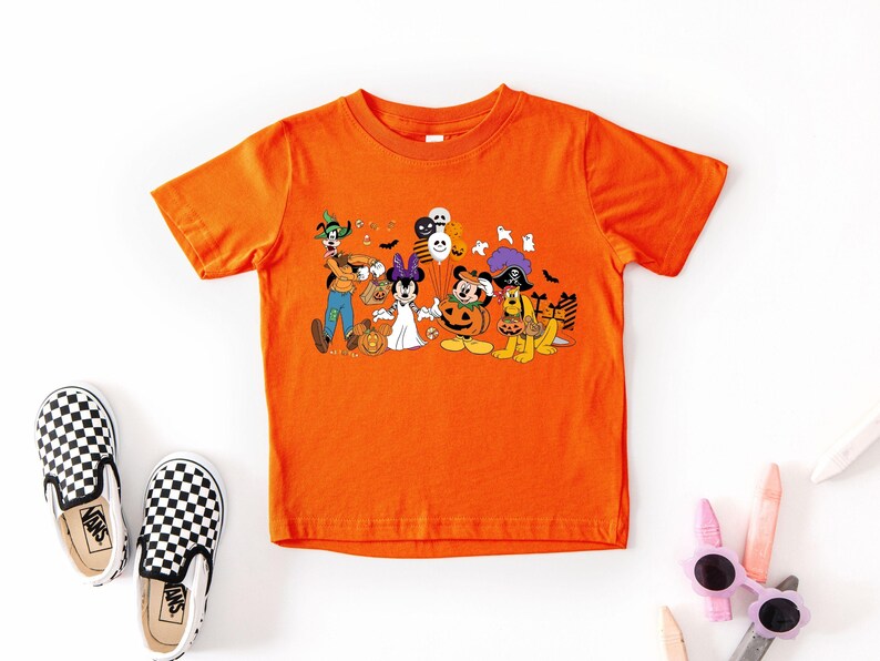May include: Orange short-sleeved t-shirt featuring a Halloween-themed graphic with cartoon characters. The graphic includes characters dressed in costumes, pumpkins, and balloons. Black and white checkered slip-on shoes are next to the shirt.