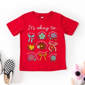 May include: A red t-shirt with the phrase "It's okay to..." and colorful illustrations. The design includes flowers, bows, and hearts, with text like "Start over" and "Have a bad day". Checkered slip-on shoes are next to the shirt.