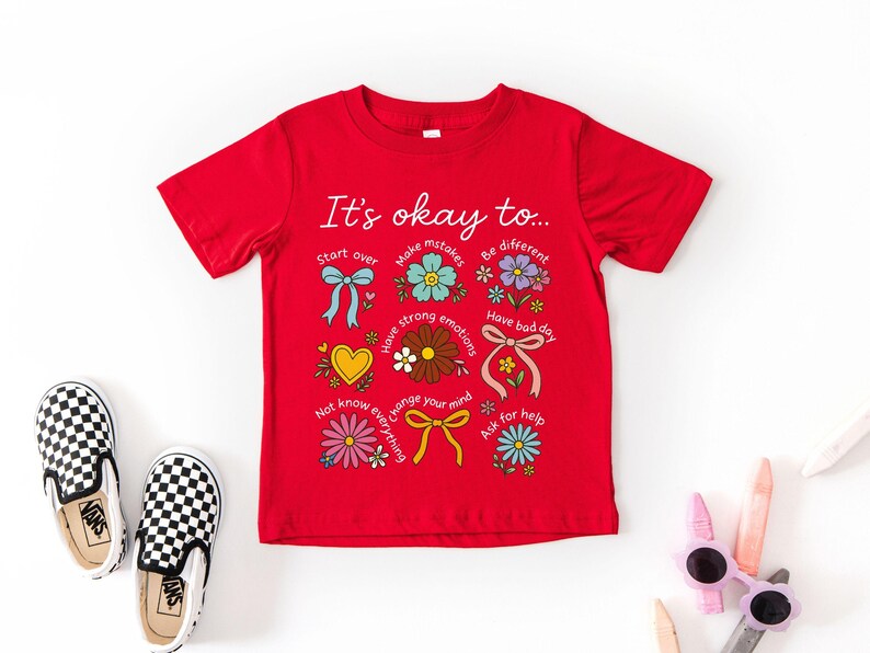 May include: A red t-shirt with the phrase "It's okay to..." and colorful illustrations. The design includes flowers, bows, and hearts, with text like "Start over" and "Have a bad day". Checkered slip-on shoes are next to the shirt.