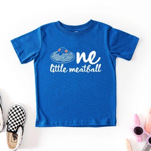 May include: A blue toddler t-shirt with the words "one little meatball" in white. The shirt features a graphic of spaghetti and meatballs. Black and white checkered slip-on shoes and sunglasses are also in the image.