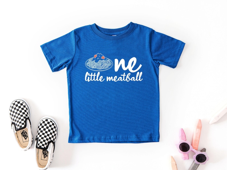 May include: A blue toddler t-shirt with the words "one little meatball" in white. The shirt features a graphic of spaghetti and meatballs. Black and white checkered slip-on shoes and sunglasses are also in the image.