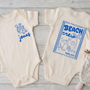 Op de afbeelding: Twee cr&egrave;mekleurige babyrompers met blauwe afbeeldingen. E&eacute;n heeft een zandemmer- en schepontwerp met de naam "jonas". De andere zegt "BEACH crew" met strandillustraties en "MIAMI, 2026". Een knuffelkonijn ligt naast de romper.