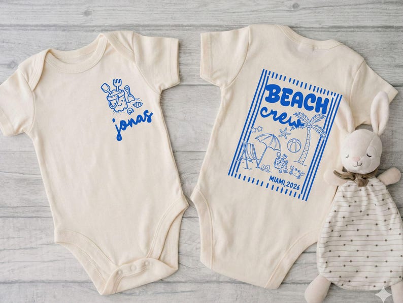 Op de afbeelding: Twee cr&egrave;mekleurige babyrompers met blauwe afbeeldingen. E&eacute;n heeft een zandemmer- en schepontwerp met de naam "jonas". De andere zegt "BEACH crew" met strandillustraties en "MIAMI, 2026". Een knuffelkonijn ligt naast de romper.