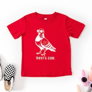 May include: A red t-shirt with a white graphic of a pigeon wearing sunglasses. The text "THAT'S COO." is below the graphic. Black and white checkered slip-on shoes are on the left. Sunglasses and crayons are on the right.