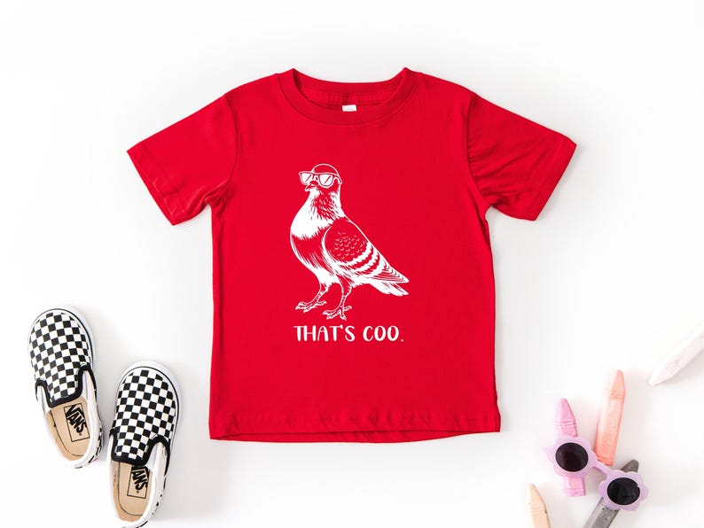 May include: A red t-shirt with a white graphic of a pigeon wearing sunglasses. The text "THAT'S COO." is below the graphic. Black and white checkered slip-on shoes are on the left. Sunglasses and crayons are on the right.