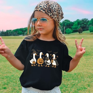 May include: Black t-shirt featuring a Halloween-themed design with four cartoon geese dressed in costumes. One goose wears a witch hat, another a pumpkin head, and others hold treats. Bats are also included in the design.