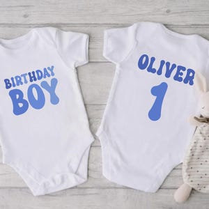 May include: Two white baby vests with blue text. One says "BIRTHDAY BOY" and the other says "OLIVER 1". A stuffed bunny with a patterned blanket is next to the second vest. The vests are likely for a first birthday.