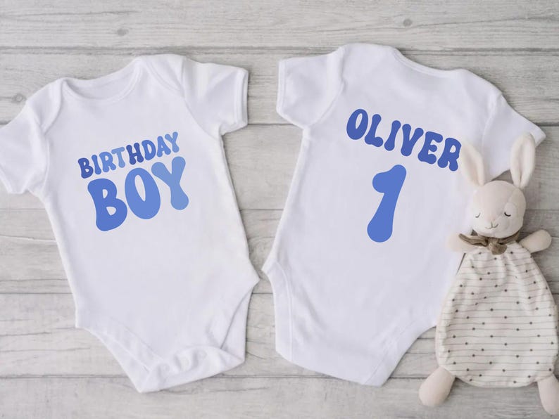 May include: Two white baby vests with blue text. One says "BIRTHDAY BOY" and the other says "OLIVER 1". A stuffed bunny with a patterned blanket is next to the second vest. The vests are likely for a first birthday.