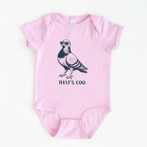 May include: A light pink baby bodysuit with a black graphic of a pigeon wearing sunglasses. The text "THAT'S COO." is printed below the bird. The bodysuit is made of soft material.
