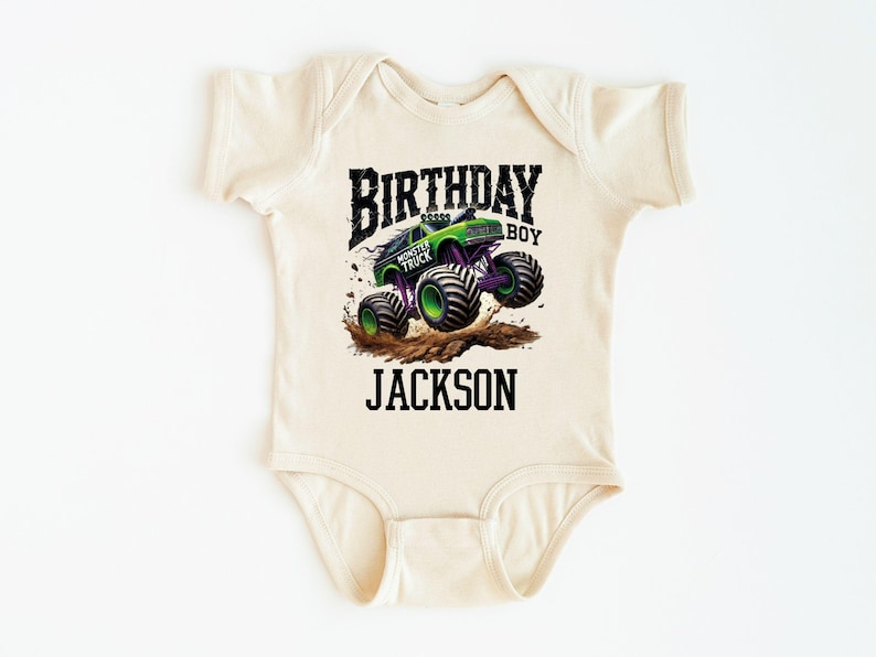 Pode incluir: Um body de beb&ecirc; bege com um gr&aacute;fico de monster truck verde e roxo. O texto diz "BIRTHDAY BOY" acima do caminh&atilde;o e "JACKSON" abaixo. O design apresenta um monster truck levantando poeira.