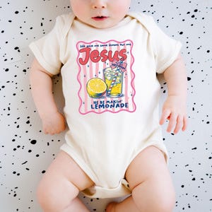 May include: Cream-colored baby onesie with a colorful graphic. The graphic features the word "Jesus" in red script, a lemon, and a glass of lemonade. Text reads "Life gave me some lemons but my Jesus he be makin' lemonade."