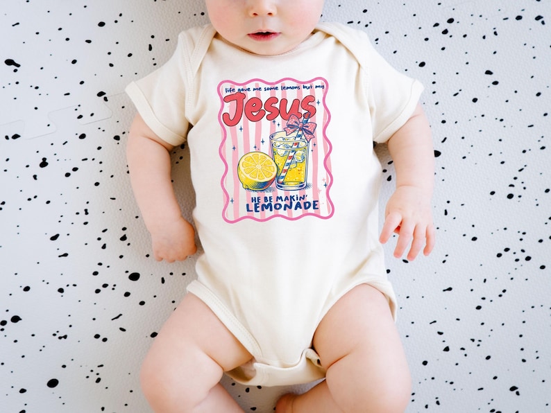 May include: Cream-colored baby onesie with a colorful graphic. The graphic features the word "Jesus" in red script, a lemon, and a glass of lemonade. Text reads "Life gave me some lemons but my Jesus he be makin' lemonade."